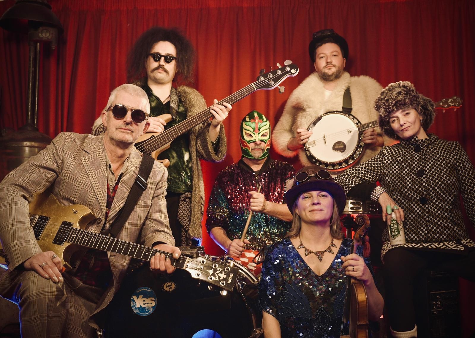 The six person band Shooglenifty posing in front of a red curtain with their various instruments